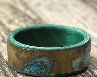 May include: A handcrafted ring with a unique design. The ring features a wide band with a green interior and a textured, bronze-colored exterior with blue accents. The ring is set against a wooden surface.