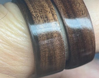 May include: Two matching rings crafted from dark brown wood with a smooth, polished finish. The rings feature a visible natural grain pattern throughout the surface, showcasing the deep brown tones and texture of the wood material in a simple band style.