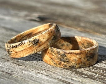 May include: Two rings crafted from light-colored wood featuring a distinct grain pattern with dark brown and black speckles. The rings have a smooth, polished finish and a rounded band profile, showcasing natural variations in the wood texture.