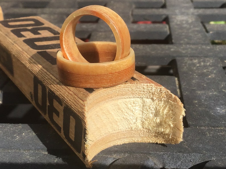 May include: Two wooden rings with a light brown and reddish-brown striped design rest on a section of a hockey stick. The rings are made of layered wood, showcasing the natural grain. The hockey stick has black text.