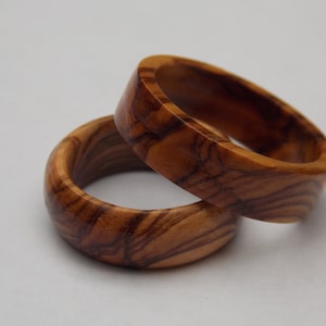 Puede incluir: Dos anillos de madera hechos de madera de olivo. Los anillos est&aacute;n apilados uno encima del otro. La veta de la madera es visible en los anillos.