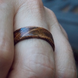 K&ouml;nnte beinhalten: Ein Holzring mit einem braunen und schwarzen Streifenmuster. Der Ring wird an einem Finger getragen.