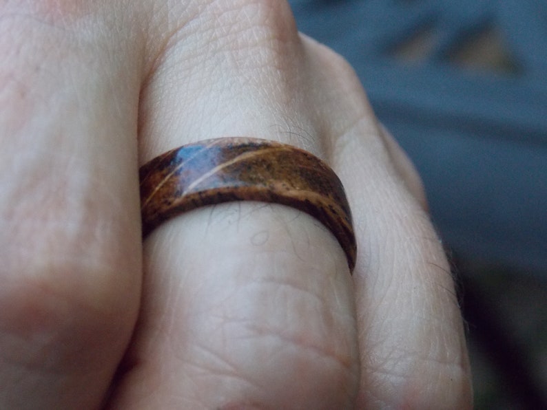 K&ouml;nnte beinhalten: Ein Holzring mit einem braunen und schwarzen Streifenmuster. Der Ring wird an einem Finger getragen.
