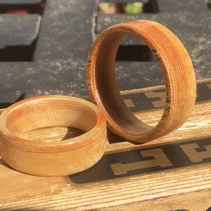 May include: Two wooden rings with a light brown and reddish-brown color scheme. The rings are made of layered wood, with visible grain patterns. One ring is larger than the other. The rings are displayed on a wooden surface.