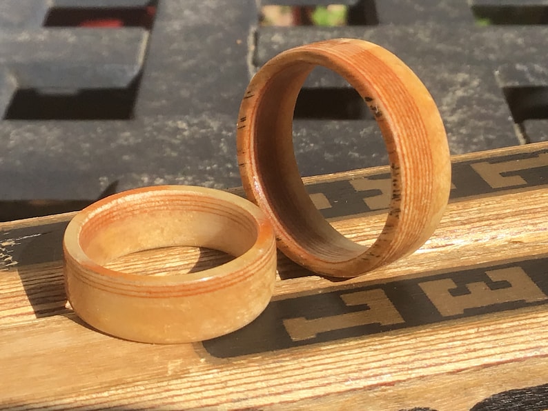 May include: Two wooden rings with a light brown and reddish-brown color scheme. The rings are made of layered wood, with visible grain patterns. One ring is larger than the other. The rings are displayed on a wooden surface.