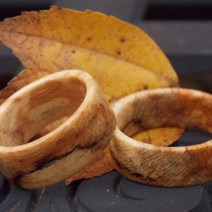 May include: Two wooden rings with a natural wood grain pattern. The rings are light brown with darker brown streaks and are set against a backdrop of a yellow leaf. The rings are likely handmade and are a unique piece of jewellery.