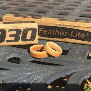 May include: Two wooden rings with a gradient of orange and tan hues sit on a dark surface. Behind them are two wooden blocks with the text "5030" and "Feather-Lite" printed in black. The rings appear to be handmade.