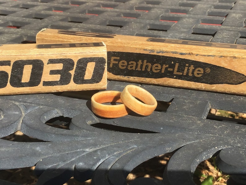 May include: Two wooden rings with a gradient of orange and tan hues sit on a dark surface. Behind them are two wooden blocks with the text "5030" and "Feather-Lite" printed in black. The rings appear to be handmade.