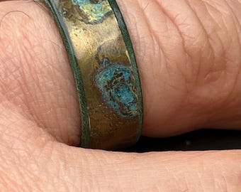 May include: A band ring featuring a metallic gold-toned surface with irregular, textured patches of oxidized turquoise and dark brown patina. The edges of the ring are finished with a dark, weathered green border, creating a distinct contrast.