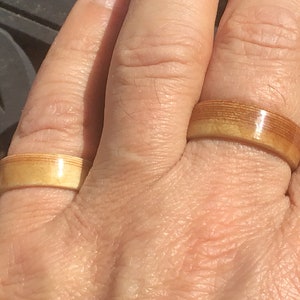 May include: Two wooden rings with a light brown color are displayed on a hand. The rings have a smooth, polished finish and a band-like design. The rings are made of layered wood, creating a unique texture.