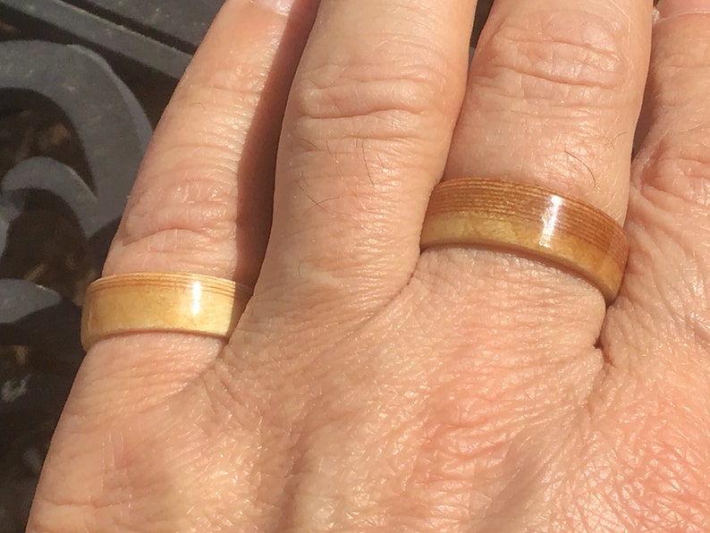 May include: Two wooden rings with a light brown color are displayed on a hand. The rings have a smooth, polished finish and a band-like design. The rings are made of layered wood, creating a unique texture.