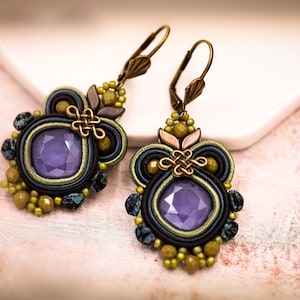 May include: A pair of handcrafted earrings featuring a central square lavender gemstone, surrounded by intricate beadwork in shades of olive green, navy, and black. The earrings have a bronze-colored metal clasp and decorative accents.