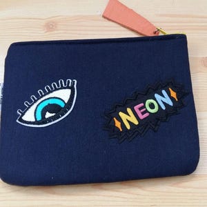 Könnte beinhalten: Marineblaue Reißverschlusstasche mit zwei aufgestickten Aufnähern: ein Auge und das Wort „NEON“ in bunten Buchstaben. Am Reißverschluss ist ein pfirsichfarbenes Etikett befestigt.