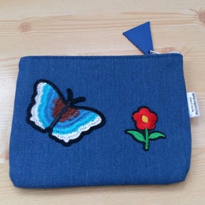 Könnte beinhalten: Eine blaue Jeans-Clutch mit einem blauen Schmetterling und einer roten Blume, die darauf gestickt sind.