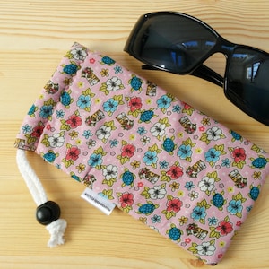 May include: Black sunglasses sit beside a pink glasses case with a drawstring closure. The case is patterned with flowers, turtles, and tiki mugs. It has a white drawstring and a black toggle. A small white tag reads "masquerade etsy.com".