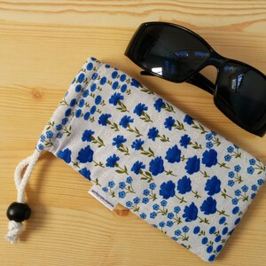 May include: A white and blue floral fabric sunglasses case with a black drawstring closure and a black toggle. The case is lying on a light wood surface next to a pair of black sunglasses.