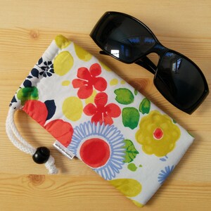 May include: A white fabric pouch with a floral pattern in shades of red, yellow, green, and blue. The pouch has a drawstring closure and a black toggle. The pouch is lying on a wooden surface next to a pair of black sunglasses.