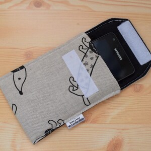 May include: A beige phone case with a black and white design of a dog. The case has a black flap with a white velcro closure. A black Samsung phone is inside the case.