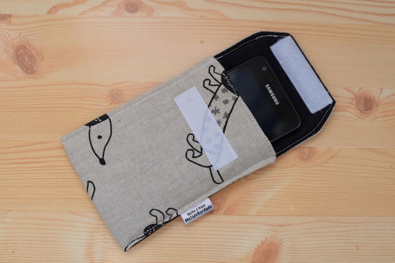 May include: A beige phone case with a black and white design of a dog. The case has a black flap with a white velcro closure. A black Samsung phone is inside the case.