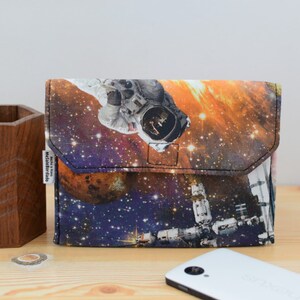 May include: A small, rectangular pouch with a zipper closure. The pouch is made of fabric and features a colorful print of an astronaut in space. The print includes stars, planets, and a space station.