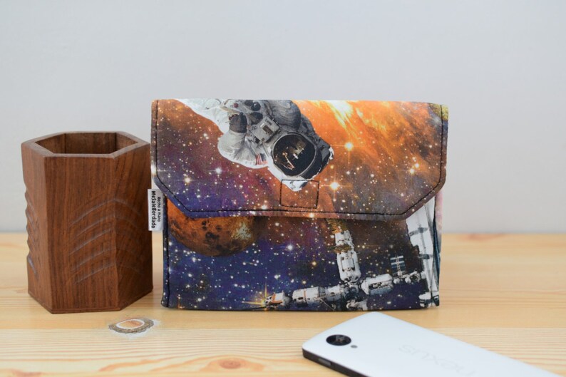 May include: A small, rectangular pouch with a zipper closure. The pouch is made of fabric and features a colorful print of an astronaut in space. The print includes stars, planets, and a space station.