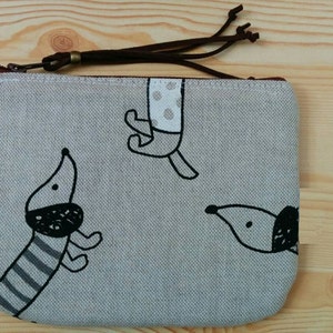 May include: A grey canvas zippered pouch with a brown leather zipper pull. The pouch features a black and white illustration of three dogs.
