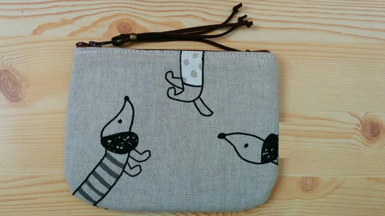May include: A grey canvas zippered pouch with a brown leather zipper pull. The pouch features a black and white illustration of three dogs.