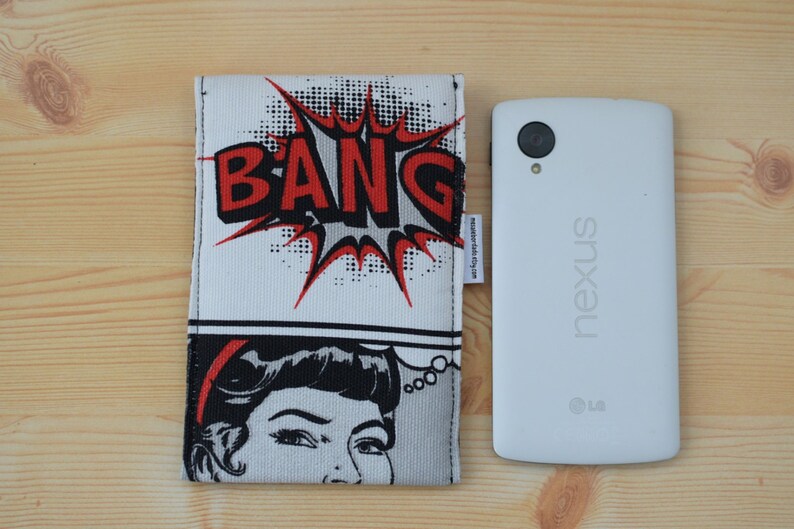 Pu&ograve; includere: Una custodia per telefono bianca con un design in stile fumetto che presenta una grafica "BANG" rossa e nera e un'illustrazione in bianco e nero di una donna. La custodia &egrave; accanto a un telefono Nexus bianco.