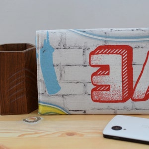 K&ouml;nnte beinhalten: Eine wei&szlig;e Stofftasche mit einem Ziegelmauermuster und roter und blauer Graffiti-Beschriftung, die "Love" sagt.