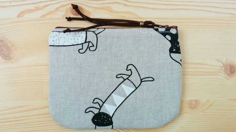 May include: A small, grey zippered pouch with a brown zipper pull. The pouch has a white and black print of stylised dogs on it.