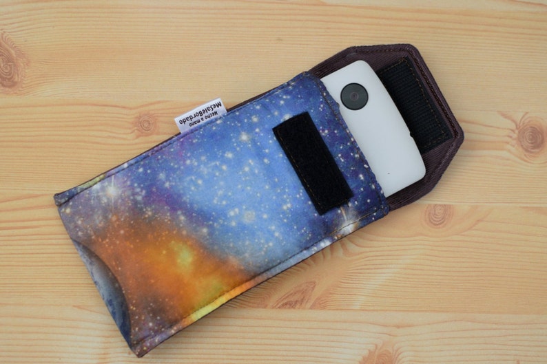 May include: A galaxy print phone case with a black strap and a white phone inside. The case has a label that reads "Made in South Africa".