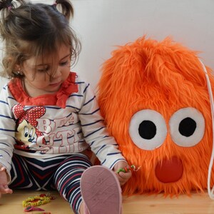May include: A young child with pigtails plays with a bright orange fuzzy drawstring bag with large white eyes and a red nose.