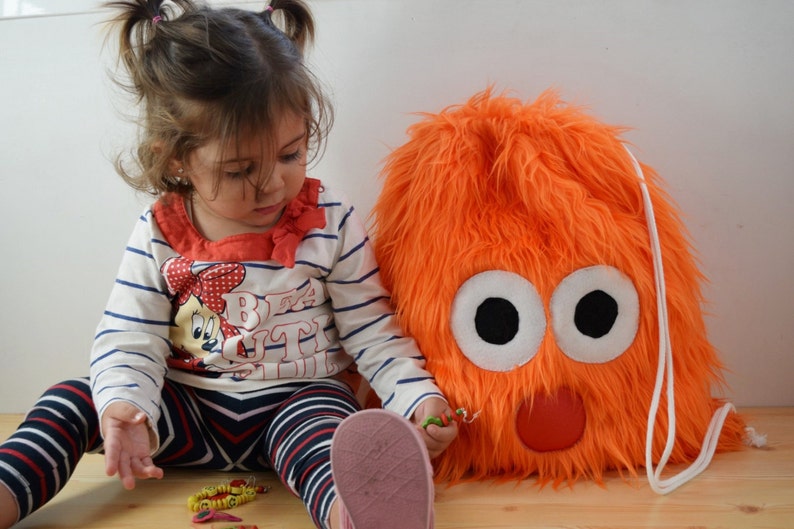 May include: A young child with pigtails plays with a bright orange fuzzy drawstring bag with large white eyes and a red nose.