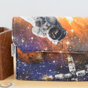 May include: A small, rectangular pouch with a zipper closure. The pouch is made of a fabric with a space-themed print featuring an astronaut, a space station, and planets. The text "Made & Sold by Melissa Bordado" is printed on a white tag attached to the pouch.