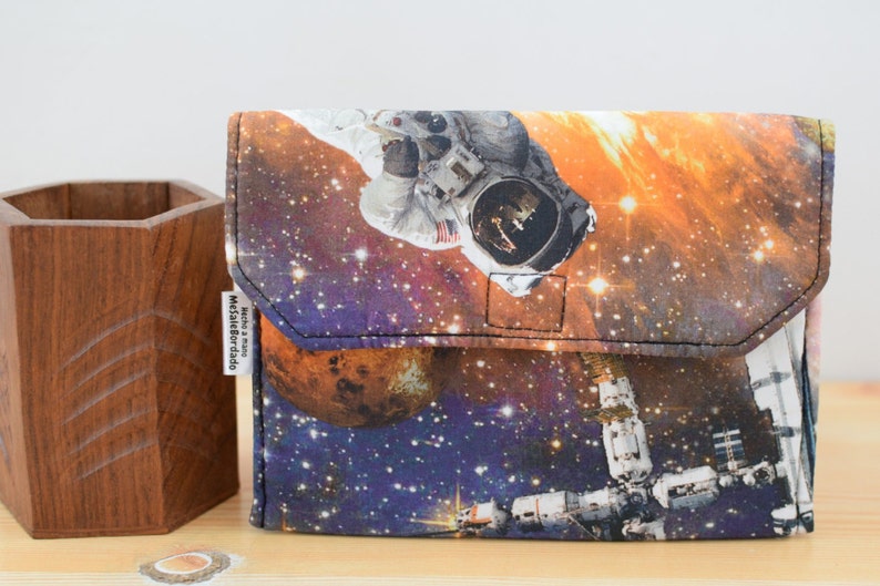 May include: A small, rectangular pouch with a zipper closure. The pouch is made of a fabric with a space-themed print featuring an astronaut, a space station, and planets. The text "Made & Sold by Melissa Bordado" is printed on a white tag attached to the pouch.