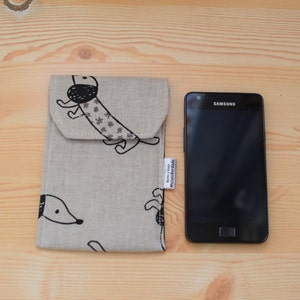 May include: A beige fabric phone case with a black and white dachshund print. The case has a flap that closes with a button. The case is next to a black Samsung smartphone.