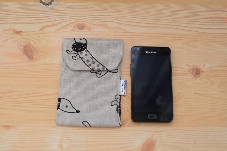 May include: A beige fabric phone case with a black and white dachshund print. The case has a flap that closes with a button. The case is next to a black Samsung smartphone.
