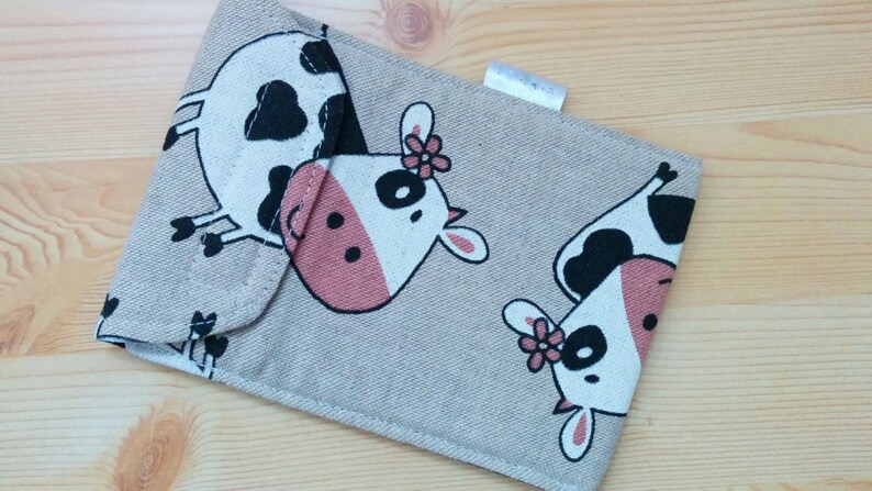 May include: A beige fabric pouch with a flap closure. The pouch is decorated with a repeating pattern of black and white cows with pink noses and black eyes. Some of the cows have a pink flower on their heads.