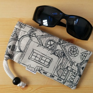 May include: A black and white patterned fabric eyeglass case with a drawstring closure and a black bead. The case is lying on a wooden surface next to a pair of black sunglasses.