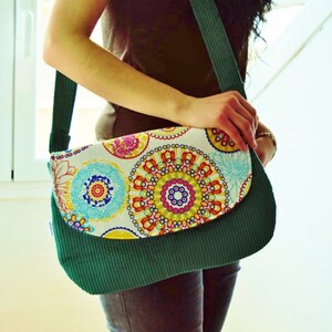 Könnte beinhalten: Eine grüne Cord-Schultertasche mit einem Klappverschluss. Die Klappe ist mit einem bunten Blumenmuster verziert, das Kreise und Wirbel zeigt.