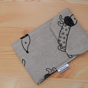 May include: A beige fabric pouch with a black and white illustration of a dog. The pouch has a label that reads "Hecho a mano /  MelafeBordado".