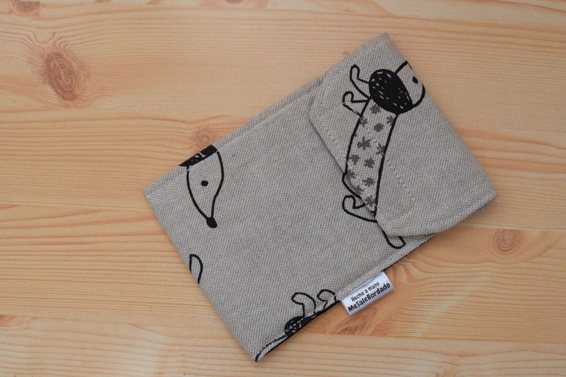 May include: A beige fabric pouch with a black and white illustration of a dog. The pouch has a label that reads "Hecho a mano /  MelafeBordado".