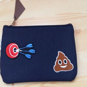 Könnte beinhalten: Eine dunkelblaue Reißverschlusstasche mit einem roten und weißen Ziel mit blauen Pfeilen und einem braunen Kack-Emoji mit einem weißen Lächeln.