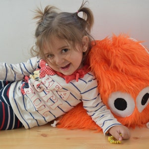 May include: A young girl with pigtails is lying on her side, leaning against a bright orange fuzzy backpack with large black eyes. She is wearing a white and blue striped shirt with the words "Big Girl" printed on it.