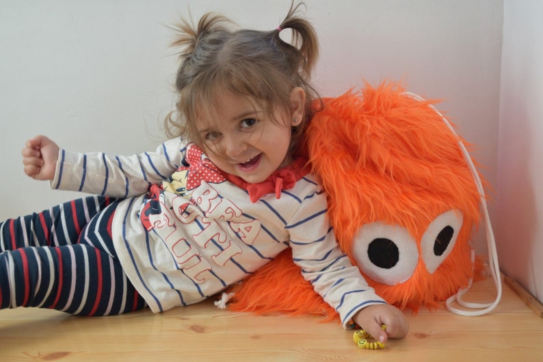 May include: A young girl with pigtails is lying on her side, leaning against a bright orange fuzzy backpack with large black eyes. She is wearing a white and blue striped shirt with the words "Big Girl" printed on it.
