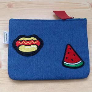 Könnte beinhalten: Eine blaue Jeans-Clutch mit einem Hotdog- und Wassermelonen-Patch. Der Hotdog-Patch ist gelb mit rotem Senf und einem braunen Brötchen. Der Wassermelonen-Patch ist rot mit grüner Schale.