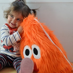 May include: A young child hugs a large orange furry toy with white eyes. The toy has a white drawstring.