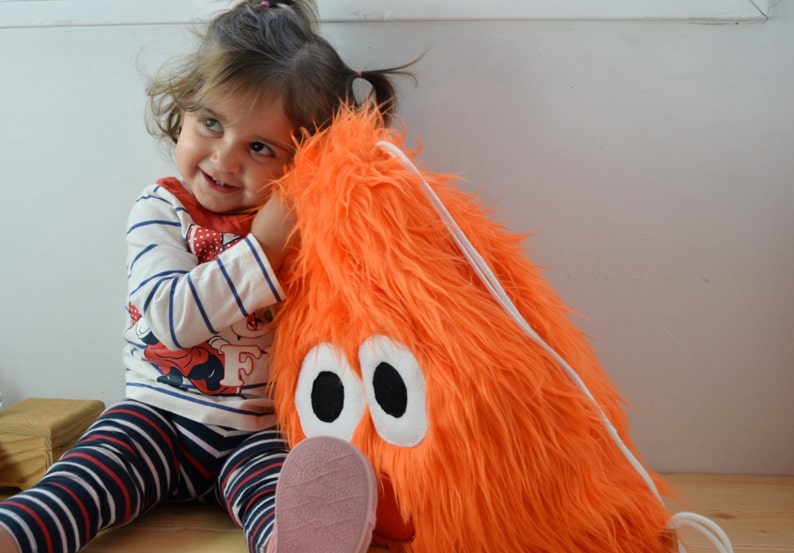 May include: A young child hugs a large orange furry toy with white eyes. The toy has a white drawstring.