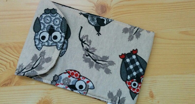 May include: A beige fabric pouch with a flap closure. The pouch is decorated with a repeating pattern of owls and leaves. The owls are in various colors, including black, white, gray, and red.