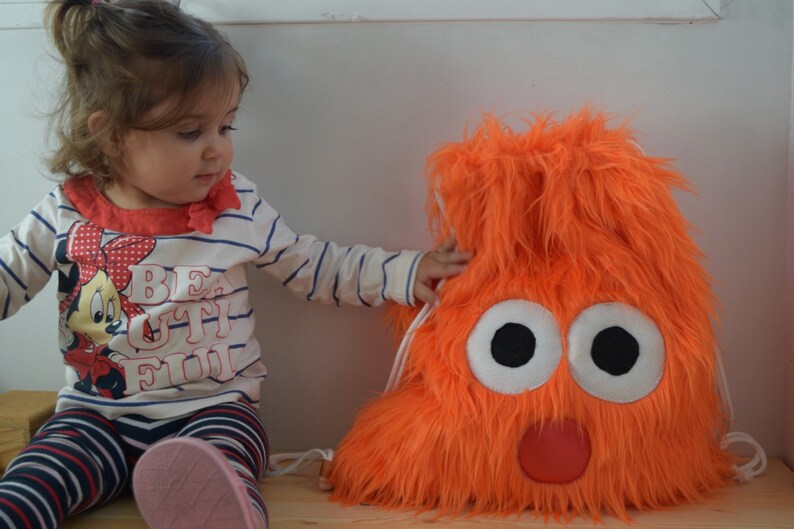 May include: A young child wearing a striped shirt with a Mickey Mouse design and the word "Beautiful" on it is looking at a bright orange furry backpack with a cartoon face. The backpack has two large white eyes with black pupils and a red mouth.